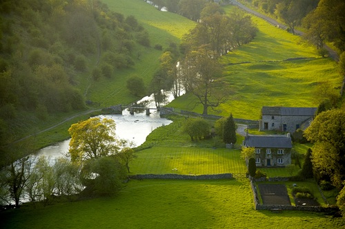 Countryside scene with a river, stone bridge, and houses surrounded by lush green fields.