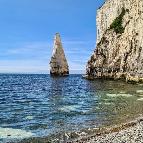 Tall sea stack near white cliffs rising from a blue ocean under a clear sky.